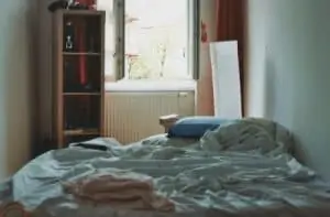 An unkempt bed beside an open window and a tall, open cabinet